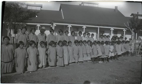 A large group of children stand in rows outside a building, all wearing similar long garments, posing for a photo in front of a porch.