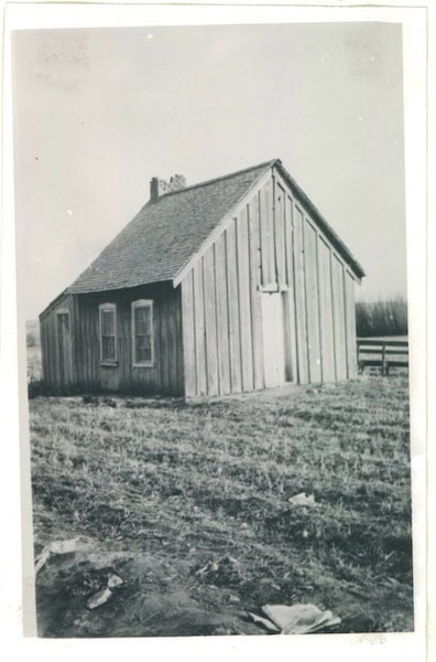 Black and white photo of a small, simple wooden house with a pitched roof, sitting on a grassy field.