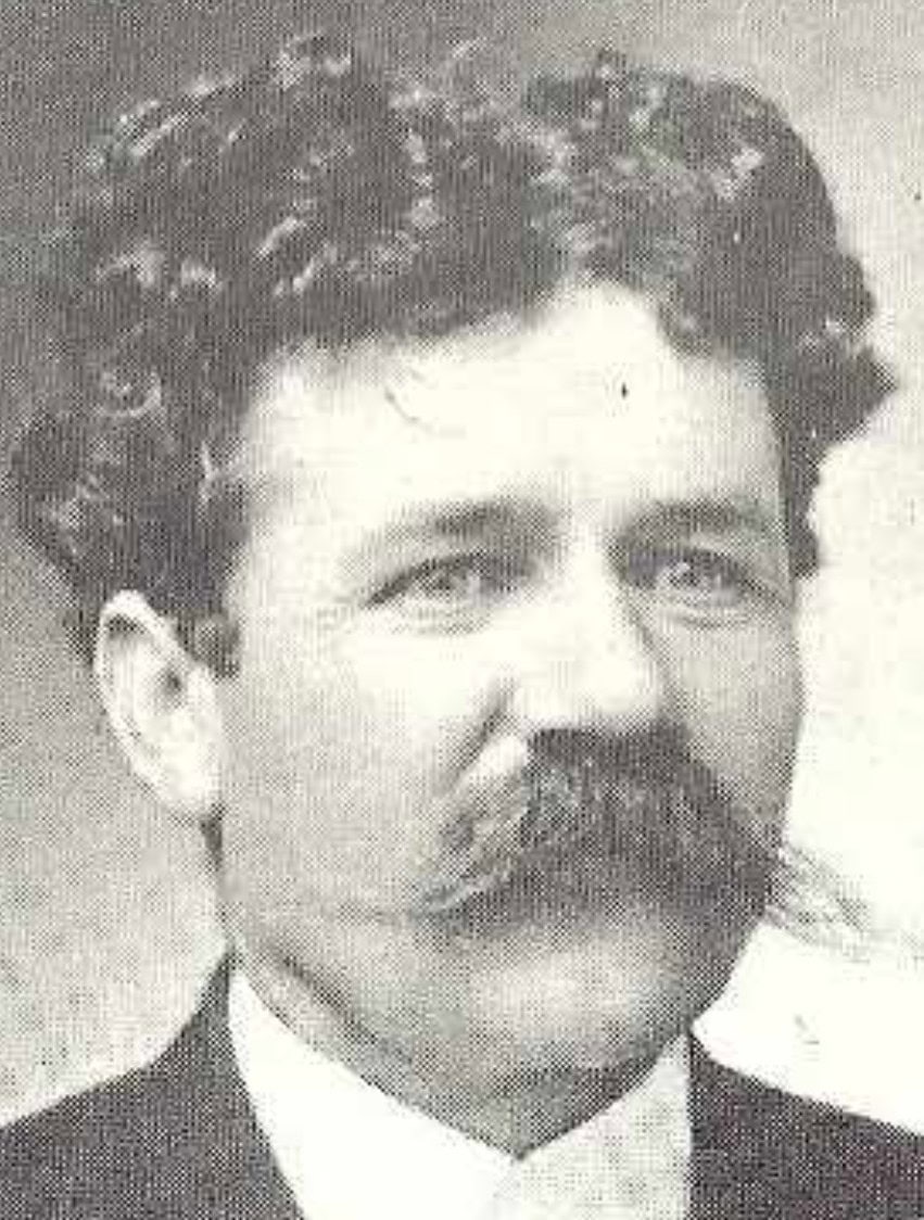 Black and white portrait of a man with curly hair, a thick mustache, and wearing a suit and tie, looking slightly off camera.