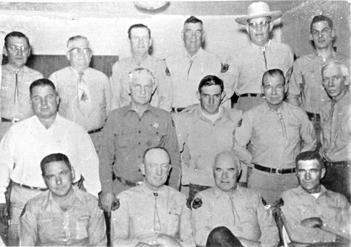 Black and white photo of sixteen men, most in uniform shirts, posed in two seated rows and two standing rows indoors.