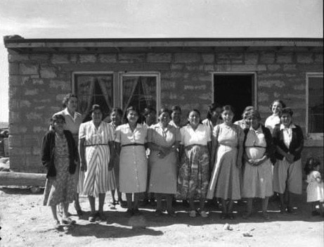 A group of women stands in front of a simple stone building on a sunny day.