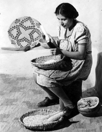 A woman sits on a low stool, husking corn into a woven basket, with other baskets and cobs around her against a plain wall.