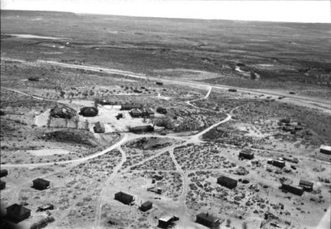 Aerial view of a sparse desert settlement with scattered small buildings, dirt roads, and minimal vegetation, surrounded by open, flat land.