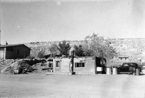 A small, single-story building with a gas pump stands near an old car and barrels in a dusty, rural landscape with sparse trees and a house in the background.