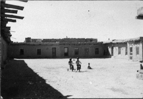 Children play in the open courtyard of a rustic adobe building complex under a clear sky. Shadows from the structures frame the scene.