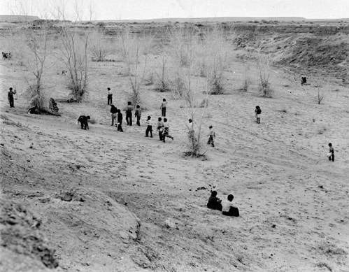 A group of people, including children, are scattered across a barren, sandy landscape with sparse vegetation and dry bushes.