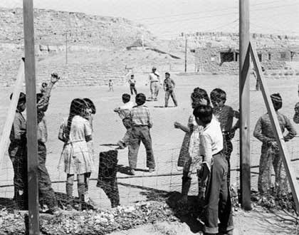 Children stand and play near a wire fence in an outdoor area with dirt ground and stone structures in the background.
