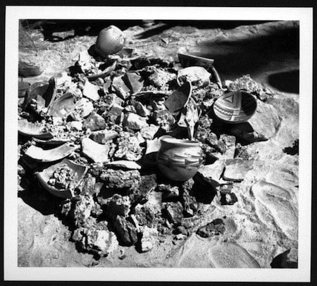 A pile of broken pottery shards and scattered rocks on sandy ground, with some pieces featuring painted designs.