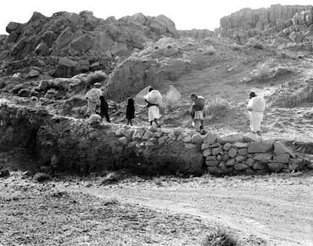 A group of people walk in a line along a rocky path with hills and large stones in the background.
