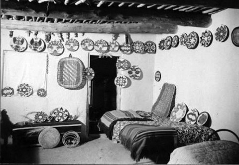A room with two beds, patterned blankets, and numerous woven baskets and trays with geometric designs displayed on the walls.