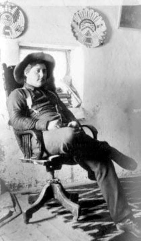 A man wearing a hat and suspenders reclines in a wooden chair, crossing his legs, with decorative plates hanging on the wall behind him.