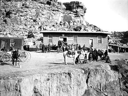 A group of people and horses are gathered outside a brick building near a rocky cliff; a wagon stands to the left.