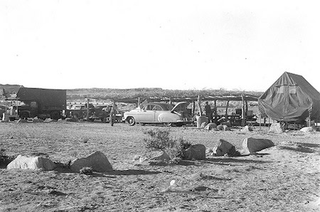Black-and-white photo of a desert campsite featuring a tent, a vintage car, a truck, and scattered rocks with sparse vegetation.