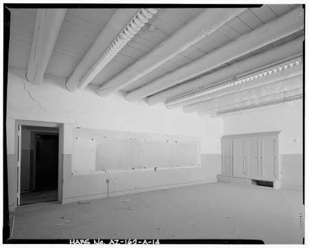 Black and white photo of an empty room with exposed ceiling beams, an open doorway, a closed cabinet, and a blank bulletin board on the wall.