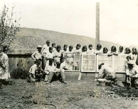 A group of children and a few adults gather outside near a wooden fence and window frames, with hills visible in the background.