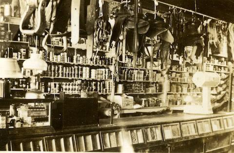 Sepia-toned photo of an old general store with shelves stocked with cans and goods, saddles hanging from the ceiling, and a scale on the counter.