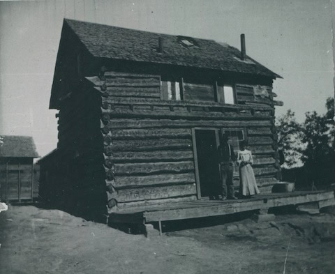 Black and white photo of two people standing in the doorway of a two-story log cabin with a slanted porch and visible wooden beams.