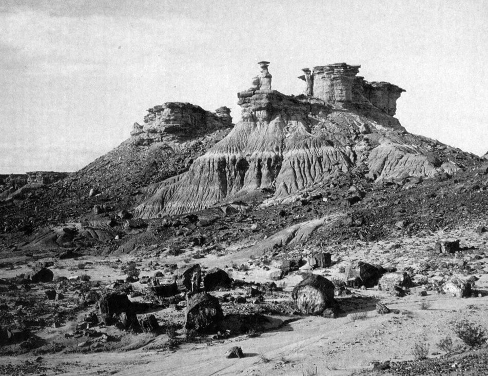 Eroded rock formations rise from a barren, rocky landscape under a clear sky, with large scattered boulders in the foreground.
