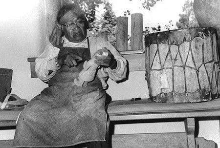 An elderly person wearing glasses sits indoors, holding and carving a wooden figure, with a large drum beside them on a bench.