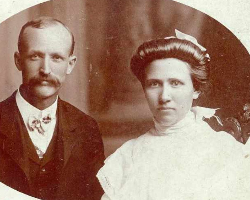 A sepia-toned portrait of a man in a suit with a mustache and a woman in a high-necked white dress, both facing the camera.
