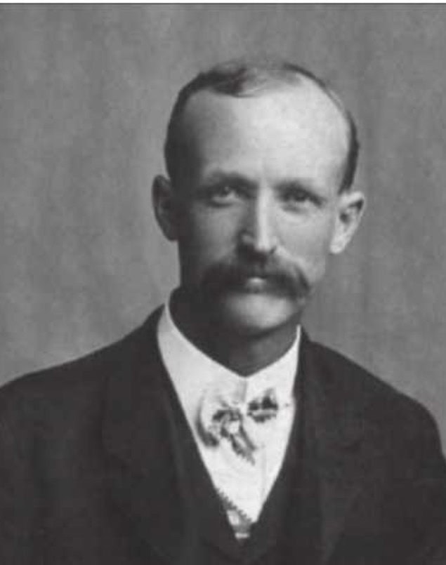 Black and white portrait of a man with a mustache, wearing a suit jacket, vest, and bow tie, facing the camera against a plain background.