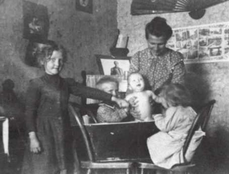 A woman bathes a baby in a tub while three children watch in a modest, old-fashioned room with pictures and a fan on the wall.
