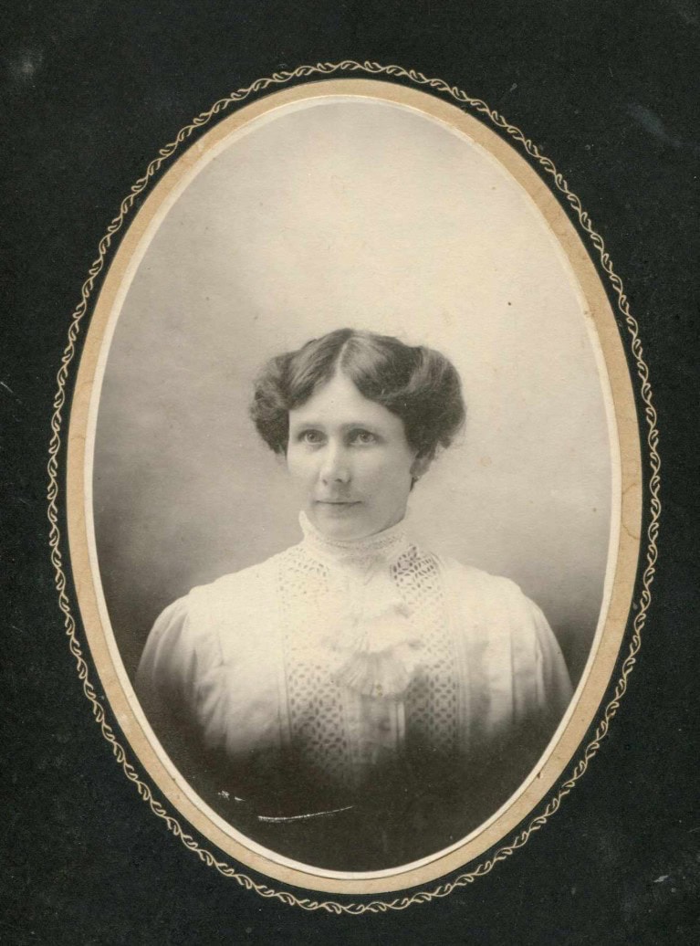 Black and white portrait of a woman with dark hair styled in waves, wearing a high-collared, lace-trimmed blouse; oval frame with decorative border.