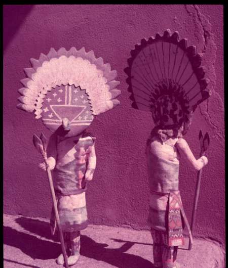Two people wearing traditional Native American ceremonial attire and large headdresses stand outdoors against a plain wall, each holding a staff.