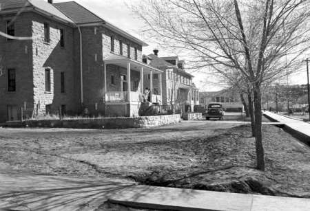 Black and white photo of two stone houses with porches, a bare tree in front, a parked car, and a sidewalk running along the street on a clear day.