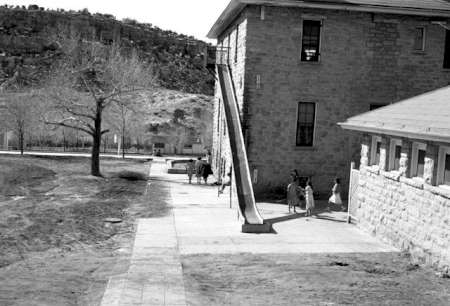 Children stand near the base of a long metal slide attached to the side of a stone building, with a barren landscape and hills in the background.