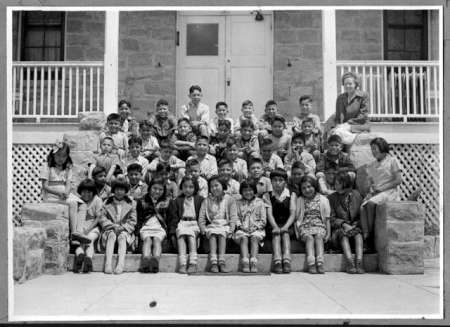 A group of school-aged children and two adults pose for a class photo on stone steps in front of a building with a porch and white railings.