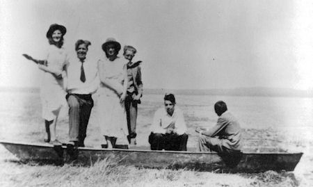 Six people pose in and around a canoe on dry land, with an open landscape and distant water visible in the background.