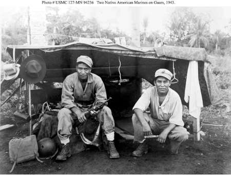 Two Native American Marines sit in front of a makeshift shelter on Guam, 1943. One Marine holds a rifle; gear and clothing are visible around them.
