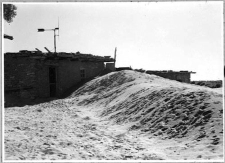 A black and white photo shows a house partially buried by a large drift of sand or dirt, with debris on the roof and a visible doorway.