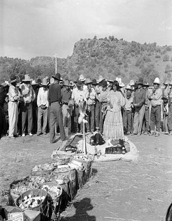 A group of people stand in a semicircle outdoors around a display of baskets and items on the ground, with hills and trees in the background.