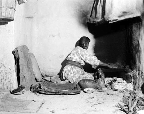 A woman kneels on the floor beside a hearth, preparing food on a flat surface in a modest, rustic room with simple household items around her.