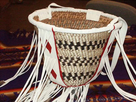 Handwoven Native American burden basket with a geometric pattern, white leather trim, and fringe, displayed on a patterned fabric background.