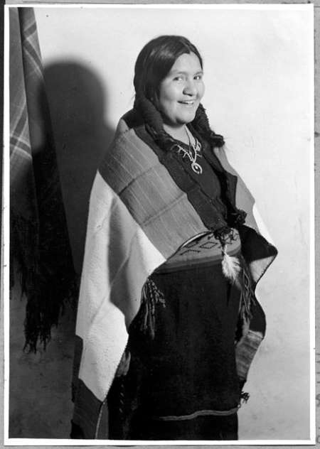 A woman in traditional clothing stands facing the camera, wearing a woven shawl and jewelry, and smiling slightly against a plain backdrop.