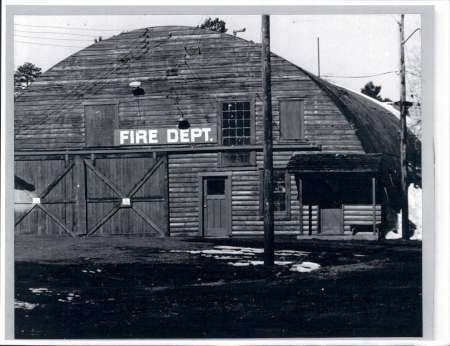 Black and white photo of a wooden, barn-like fire department building with large doors and a "FIRE DEPT." sign above the entrance.