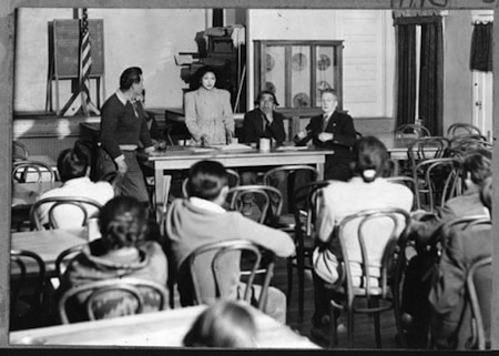 A group of people sit and listen to four individuals speaking at a table in a classroom or meeting room setting.