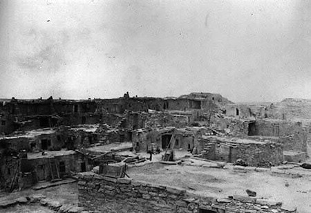 Black and white photo of an ancient Pueblo village with stone and adobe structures under a cloudy sky.