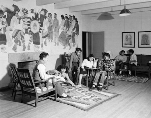 A group of people sit and converse in a room with a mural on the wall, wooden chairs, a couch, and framed artwork.