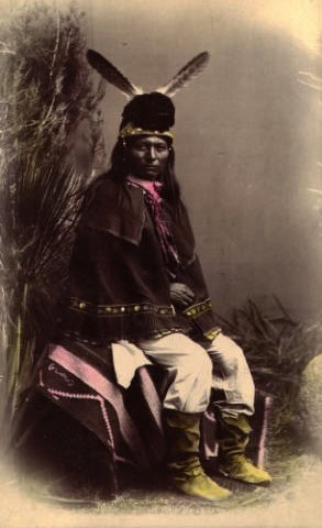 A Native American man sits on a patterned blanket, wearing traditional clothing, boots, and a headdress with two upright feathers. He poses in front of a naturalistic backdrop.