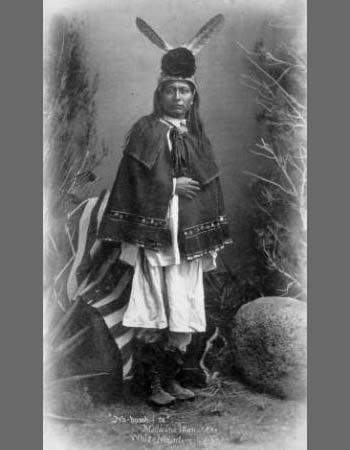 A Native American man stands in traditional attire with feathers in his hair, posing for a black-and-white studio portrait, surrounded by plants and rocks.