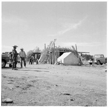 Black-and-white photo of a rural scene with people walking near a simple wood structure, a canvas tent, stacked branches, and several vehicles parked nearby.