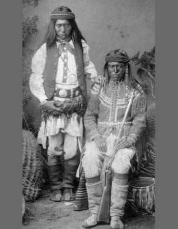 Two Indigenous men in traditional clothing pose for a historical black-and-white portrait; one is standing, and the other is seated holding a rifle.