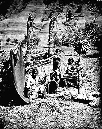 Four people sit and stand beside a woven shelter and tree in a rural outdoor setting, with hills in the background.