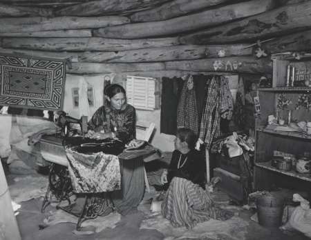 Two women sit inside a log cabin, one sewing at a machine and the other sitting on the floor; fabric, clothing, and household items fill the small, rustic room.