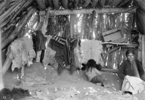 Two children sit inside a rustic wooden shelter with blankets and animal hides hanging on the wall; sunlight filters through gaps in the structure.