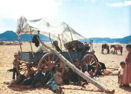 A group of people sit and rest under a covered wooden wagon in a dry, open landscape with horses and mountains in the background.